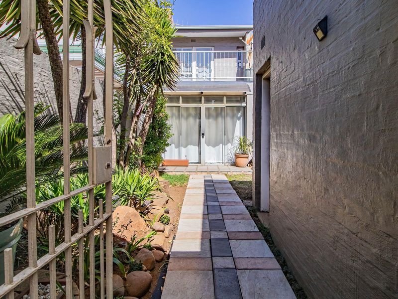 A rear exterior view of the townhouse featuring a paved walkway leading to sliding glass doors, a small garden area, and an upper-level balcony.