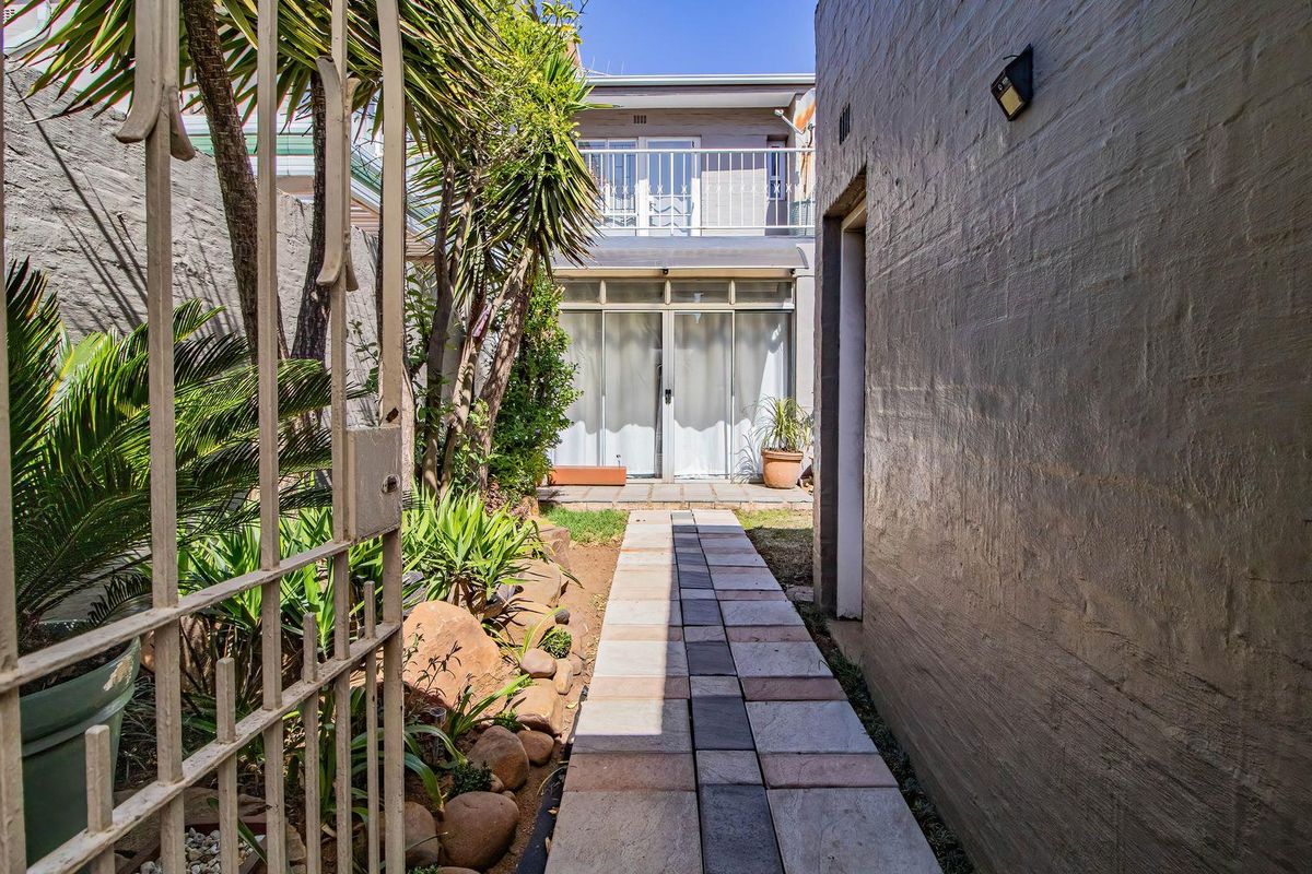A rear exterior view of the townhouse featuring a paved walkway leading to sliding glass doors, a small garden area, and an upper-level balcony.