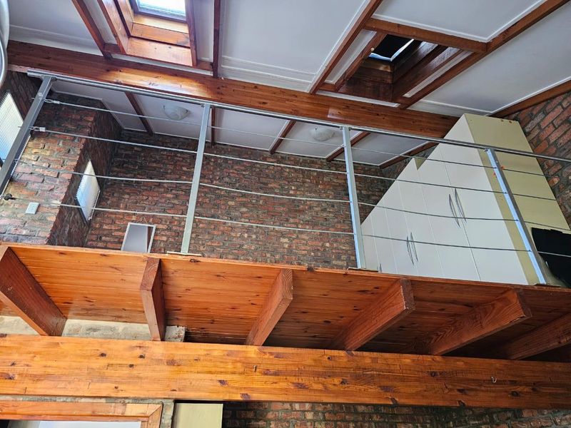 A mezzanine bedroom level featuring exposed brick walls, timber beams, skylights, and a modern stainless steel cable balustrade with built-in storage.