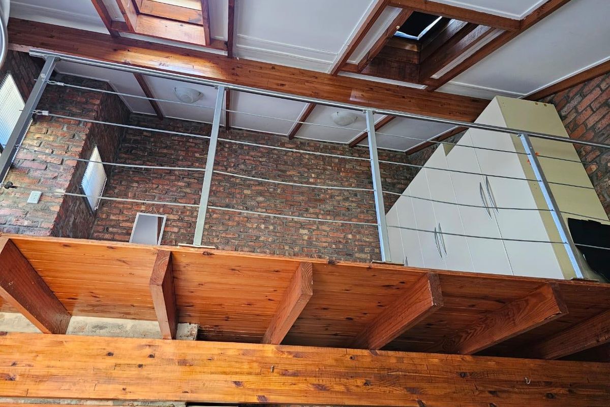 A mezzanine bedroom level featuring exposed brick walls, timber beams, skylights, and a modern stainless steel cable balustrade with built-in storage.