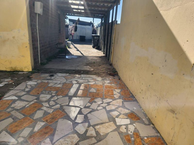 A narrow rear exterior walkway featuring a paved path and a covered structure, showing signs of wear and requiring maintenance.