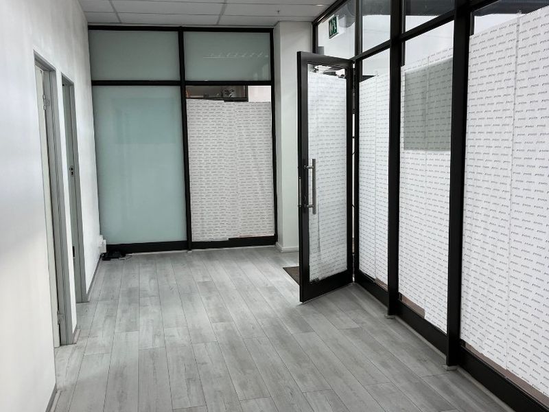 A bright, modern commercial hallway featuring laminate flooring, suspended ceiling, and glass partition walls with aluminium framing.