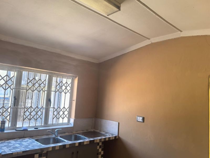 A functional kitchen area featuring a double bowl sink, tiled countertop, and security bars on the window, currently requiring cosmetic finishing and painting.