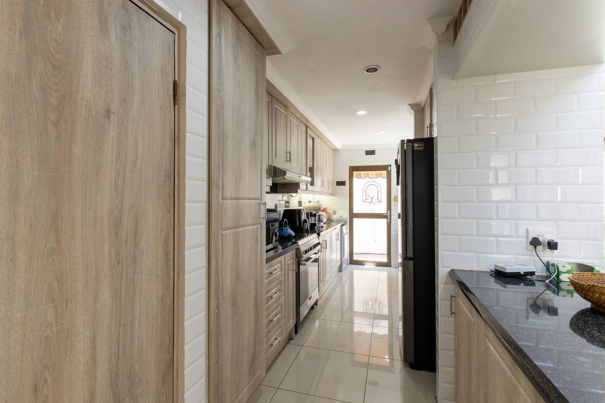 A galley-style kitchen featuring light wood-grain cabinetry, black granite countertops, white subway tile backsplash, and polished floor tiles leading to an exterior door.