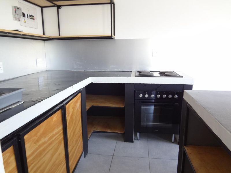 A compact kitchen featuring a built-in oven, electric hob, metal splashback, and open-style cabinetry with tiled flooring.