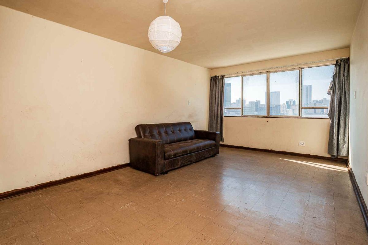 A spacious living area featuring large windows with city views, vinyl tile flooring, and neutral walls in need of cosmetic refreshment.