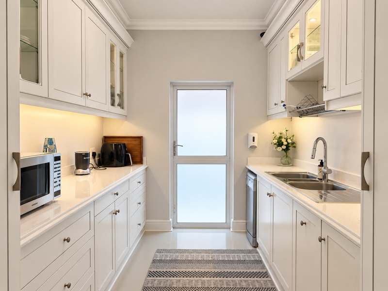 A modern, well-appointed scullery featuring crisp white cabinetry, quartz countertops, under-cabinet lighting, and a double stainless steel sink with a frosted glass exterior door.