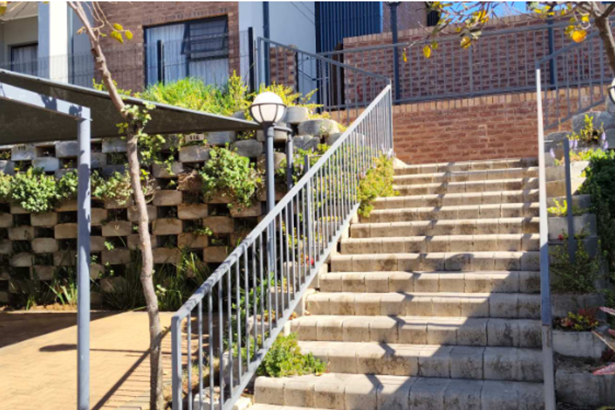 Exterior view of a multi-level apartment complex featuring a paved staircase, concrete block retaining walls, and a shaded parking area.