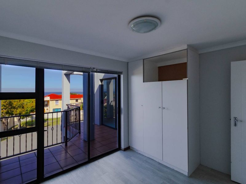 A bright, empty bedroom featuring built-in wardrobes, laminate flooring, and a sliding glass door leading to a private balcony with neighborhood views.