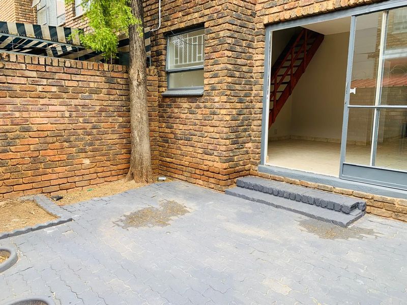 A rear exterior view of an apartment featuring a brick facade, a paved patio area, and a sliding glass door leading to the interior staircase.