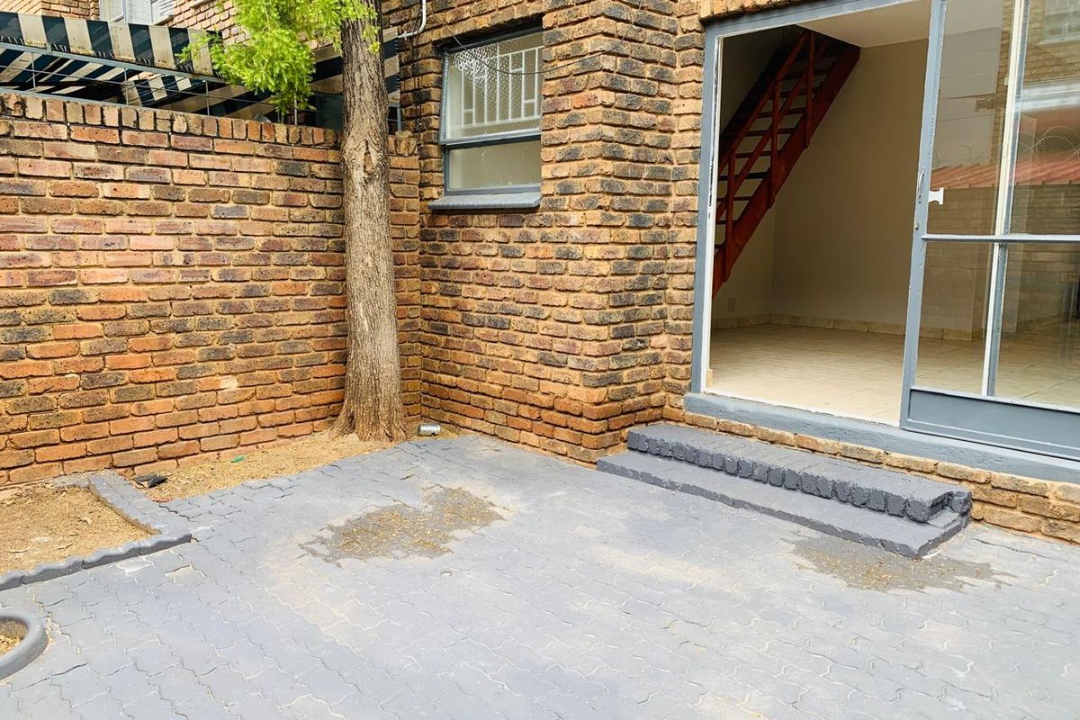 A rear exterior view of an apartment featuring a brick facade, a paved patio area, and a sliding glass door leading to the interior staircase.