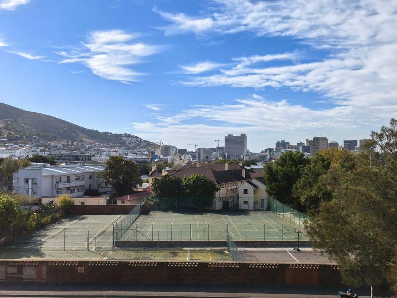 Elevated urban view overlooking a tennis court facility with surrounding residential buildings and mountain backdrop.