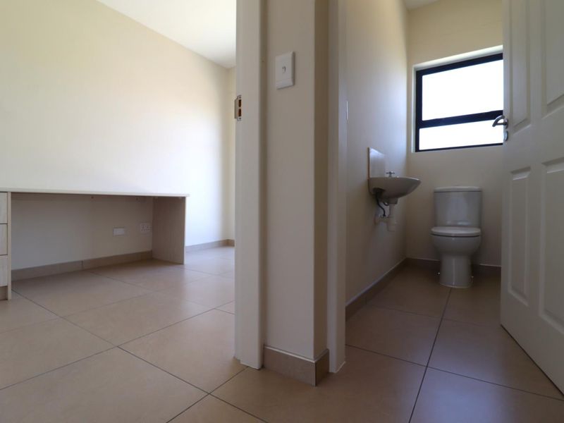 A functional bathroom featuring a wall-mounted basin and toilet, adjacent to a small room with a built-in desk area.