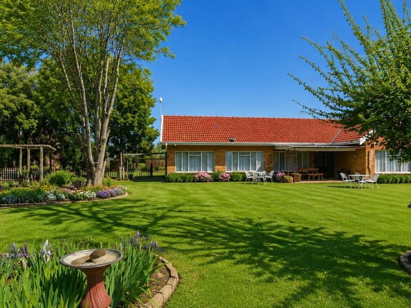 A well-maintained rear garden featuring a lush green lawn, mature trees, landscaped flower beds, and a brick house with a terracotta tiled roof.