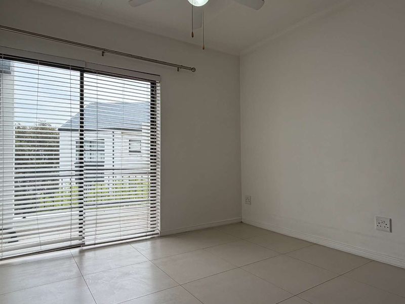 A bright, empty bedroom featuring neutral tiled flooring, a ceiling fan, and a large sliding glass door with fitted venetian blinds.