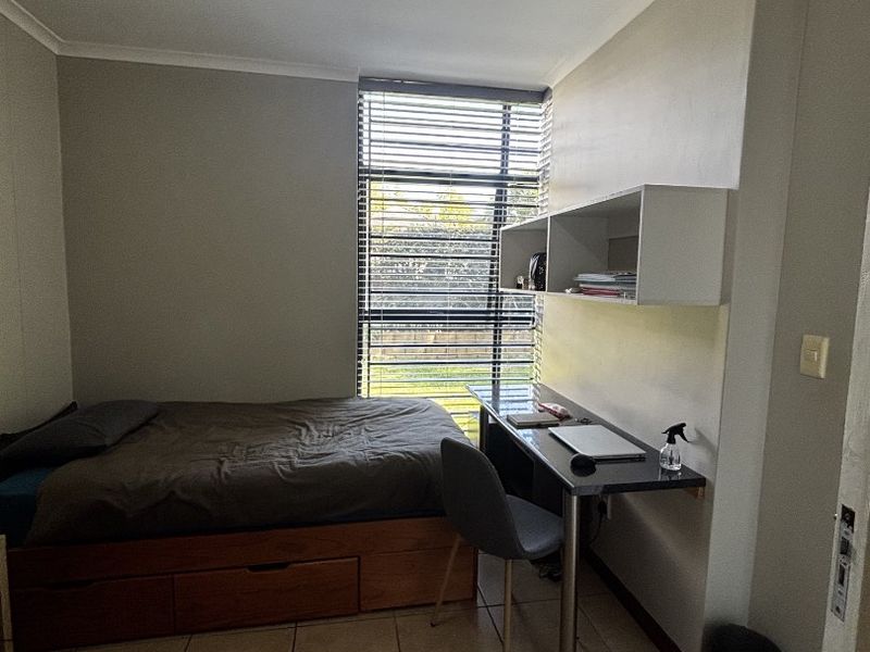 A functional bedroom featuring tiled flooring, a built-in desk with overhead shelving, and large windows with venetian blinds.