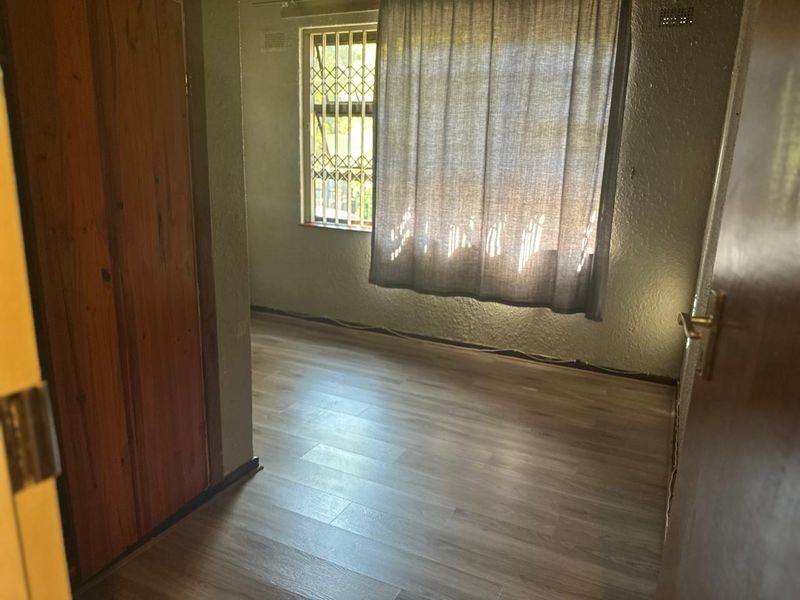 An empty bedroom featuring laminate flooring, a built-in wooden cupboard, and a window fitted with security bars.
