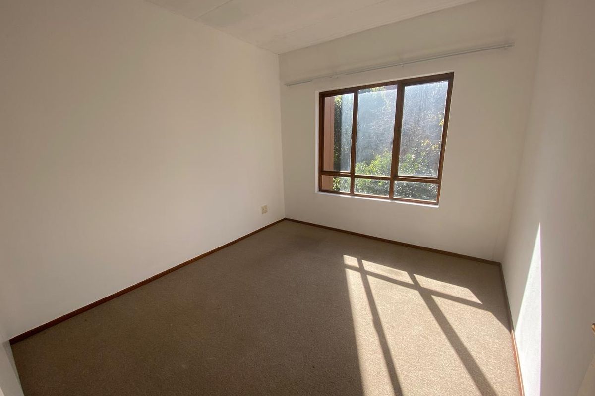A bright, empty bedroom featuring neutral walls, carpeted flooring, and a large window providing natural light and a view of the outdoors.