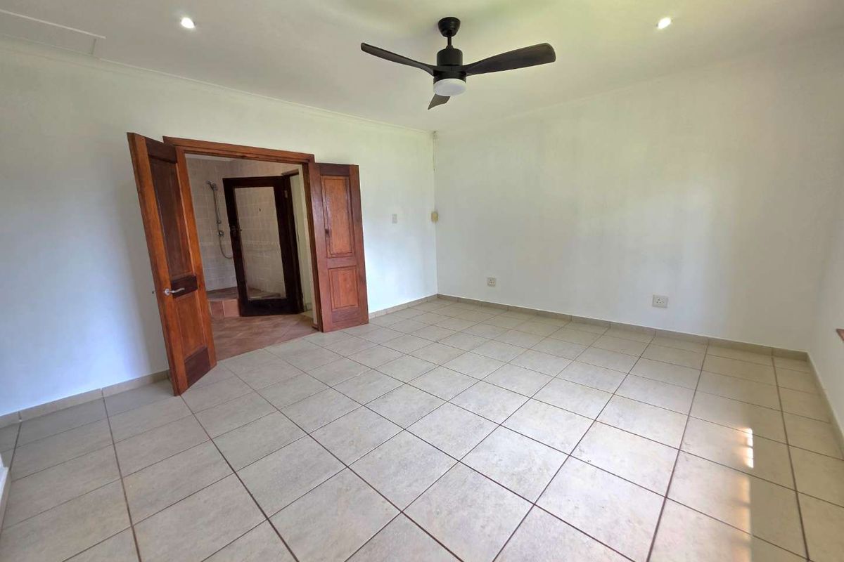 A spacious, empty bedroom featuring neutral tiled flooring, a ceiling fan, recessed lighting, and double wooden doors leading to an en-suite bathroom.