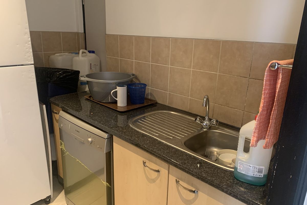 A functional kitchenette area featuring a stainless steel sink, dark granite-style countertop, light wood cabinetry, and tiled splashback.