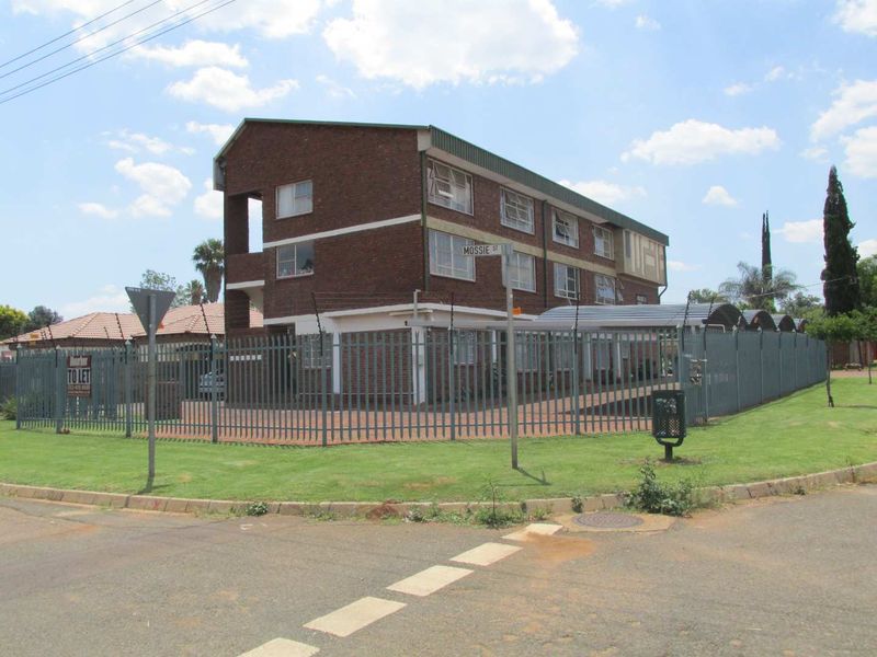 Exterior view of a multi-story brick apartment building featuring secure perimeter fencing, a paved driveway, and covered carport parking.