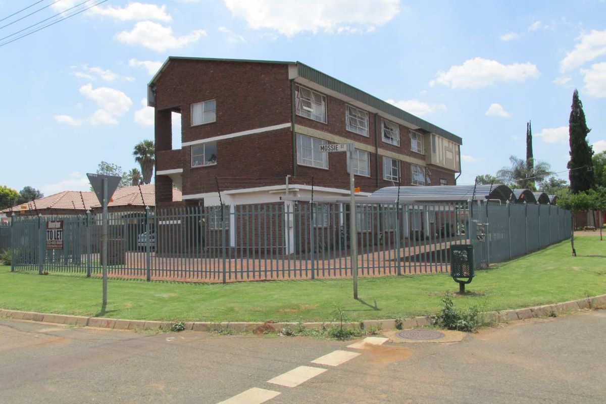 Exterior view of a multi-story brick apartment building featuring secure perimeter fencing, a paved driveway, and covered carport parking.