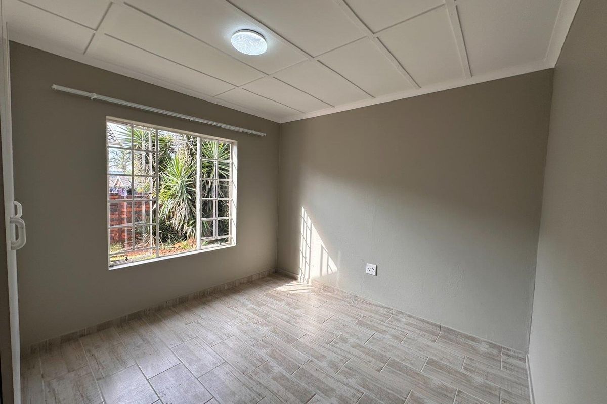 A bright, empty bedroom featuring neutral wall tones, tiled flooring, and a large window with security bars.