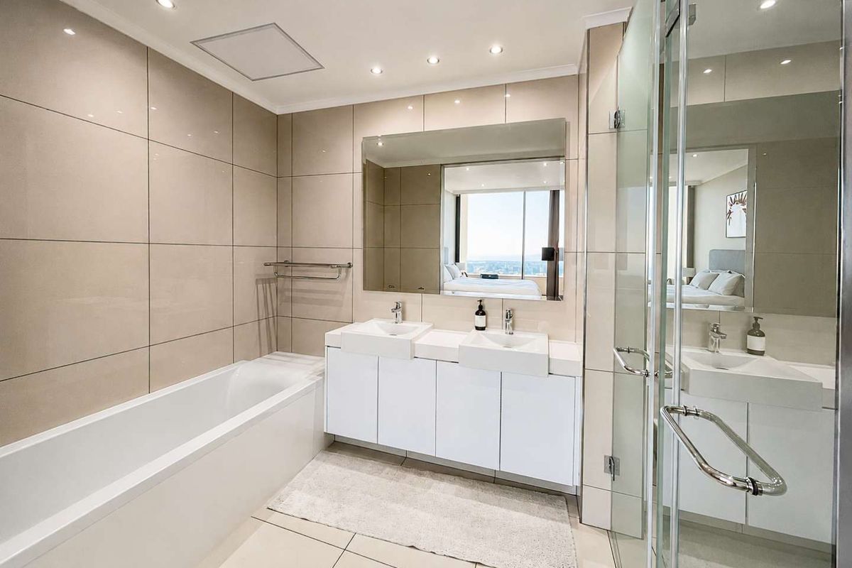 Modern bathroom featuring a double vanity, built-in bathtub, and a separate glass-enclosed shower with neutral floor-to-ceiling tiling and recessed lighting.