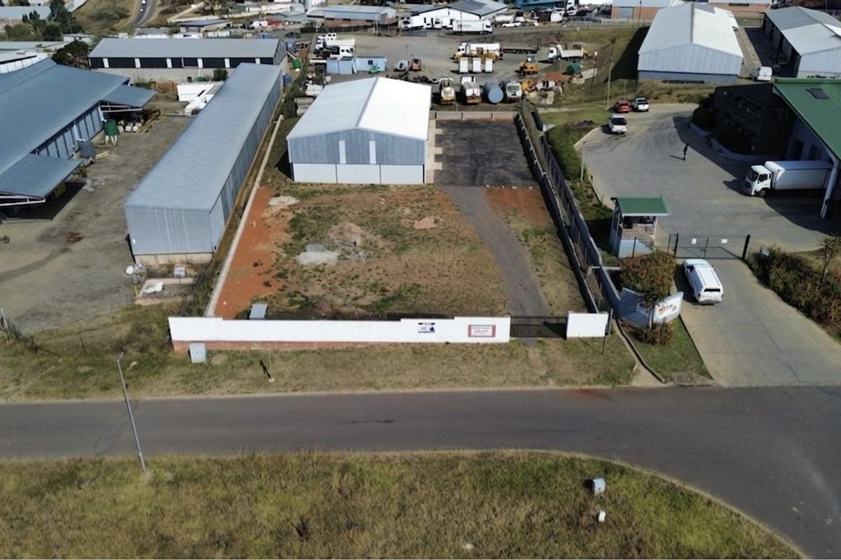 Aerial view of a commercial vacant land plot featuring a perimeter wall, security gate, guard hut, and an existing metal warehouse structure.