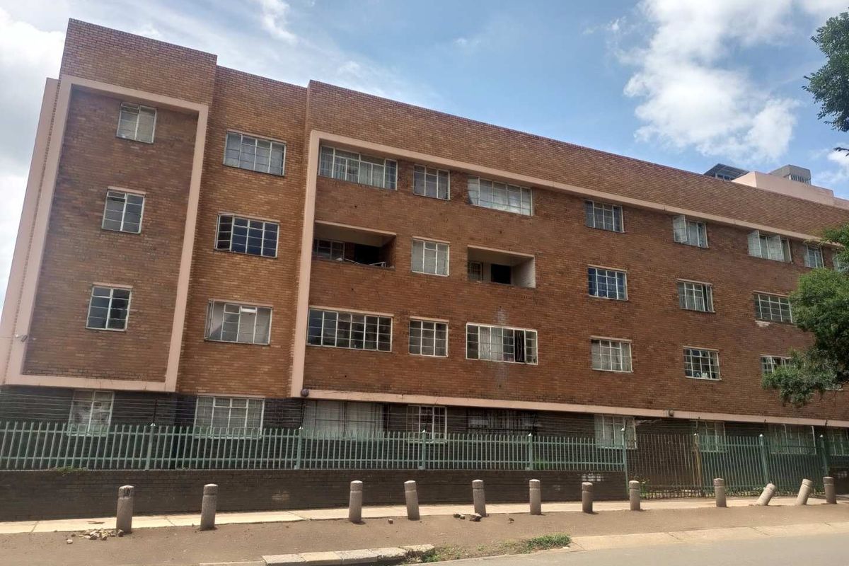 Exterior view of a multi-story brick apartment building featuring steel-framed windows and perimeter security fencing along the street frontage.