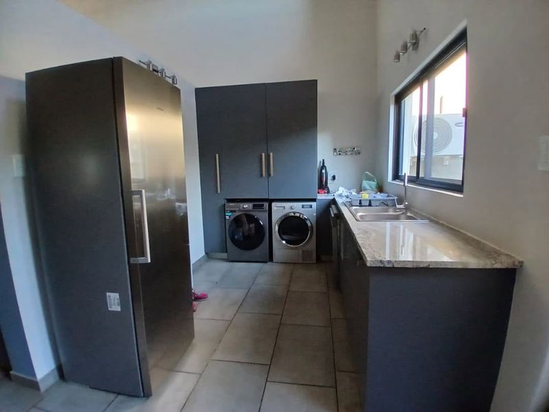 A functional laundry room featuring modern dark cabinetry, stone countertops, a stainless steel sink, and tiled flooring with space for laundry appliances.