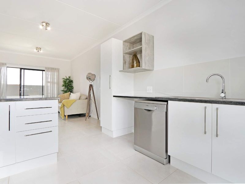 Modern open-plan kitchen and living area featuring white cabinetry, dark laminate countertops, tiled flooring, and direct access to a balcony through sliding glass doors.