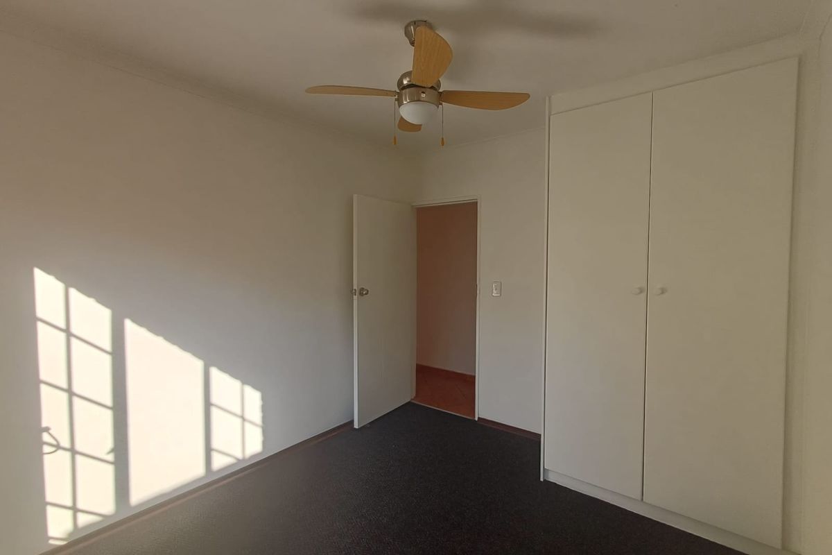 A clean, empty bedroom featuring neutral walls, dark carpeting, a built-in wardrobe, and a ceiling fan.