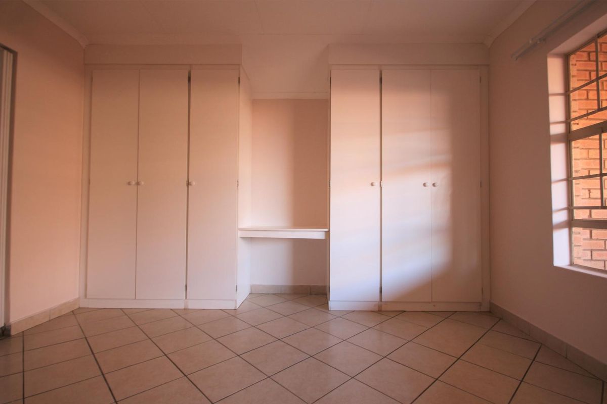A bright, empty bedroom featuring neutral tiled flooring and extensive built-in cupboard storage with an integrated desk area.