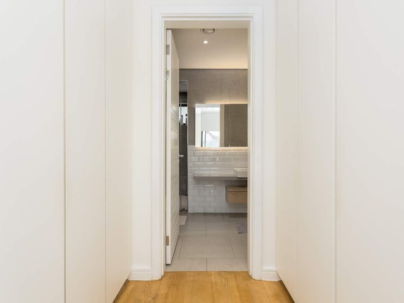 A clean, modern hallway featuring built-in cupboards and timber flooring, leading directly into a contemporary bathroom with tiled finishes.