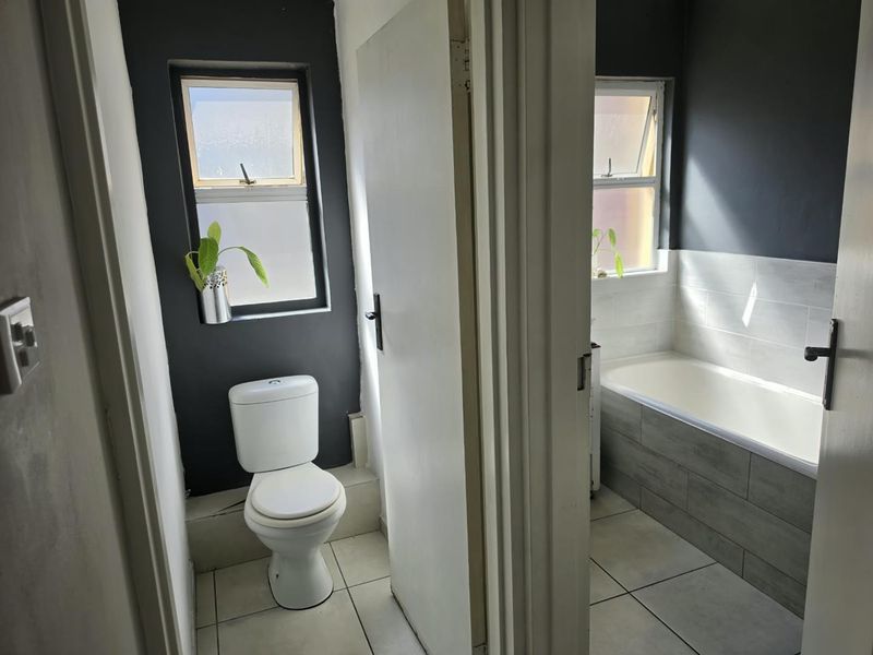 A functional bathroom featuring a separate toilet and bathtub area with tiled finishes and natural light from casement windows.
