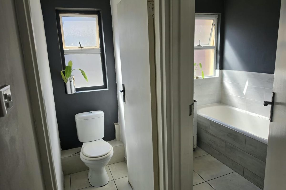 A functional bathroom featuring a separate toilet and bathtub area with tiled finishes and natural light from casement windows.