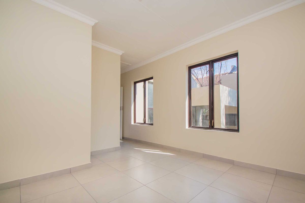 A bright, empty living area featuring neutral tiled flooring, cream-colored walls, crown molding, and dark-framed windows providing natural light.