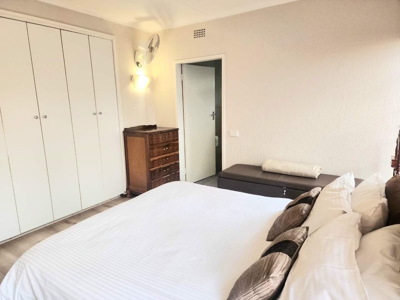 A well-lit bedroom featuring built-in cupboards, laminate flooring, and a wall-mounted fan, providing a functional and comfortable living space.