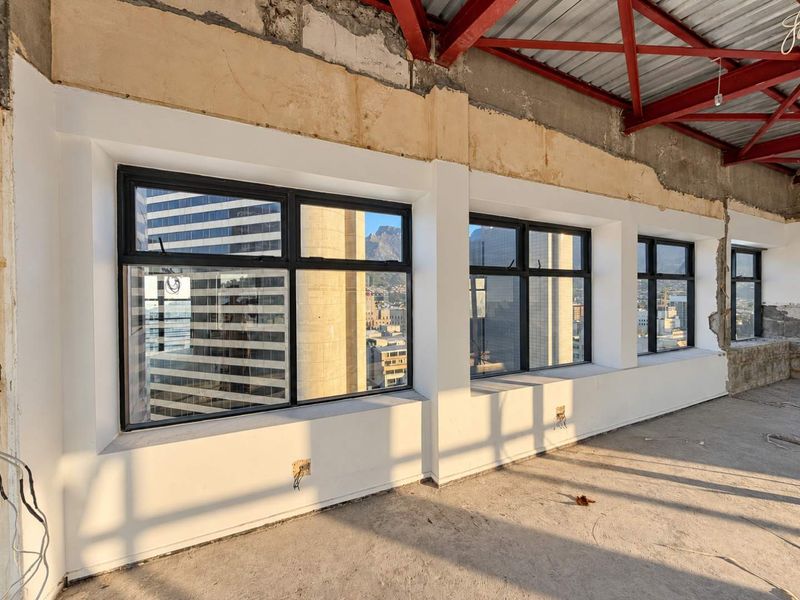 An unfinished open-plan penthouse space featuring exposed steel roof trusses, concrete flooring, and large black aluminium windows with city views.