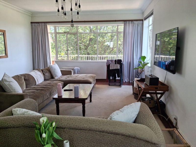 A spacious living room featuring large windows that provide ample natural light, neutral carpeting, and classic ceiling cornicing.