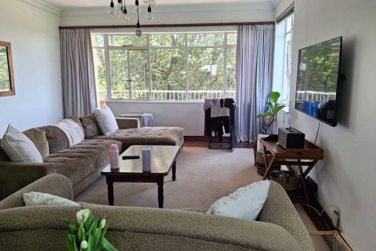 A spacious living room featuring large windows that provide ample natural light, neutral carpeting, and classic ceiling cornicing.