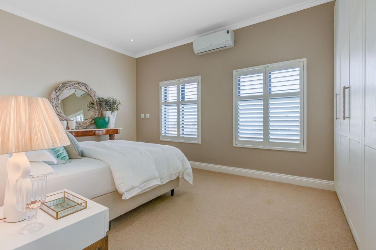A well-maintained bedroom featuring neutral carpeting, built-in wardrobes, plantation shutters, and a wall-mounted air conditioning unit.