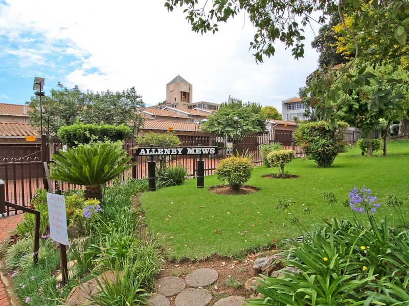 A well-maintained front entrance and garden area of the Allenby Mews townhouse complex, featuring established landscaping, brick pathways, and secure perimeter fencing.