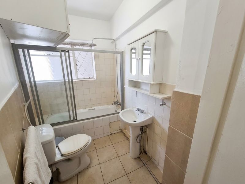 A dated bathroom featuring a bathtub with a shower screen, pedestal basin, and toilet, requiring cosmetic updates to modernize the space.