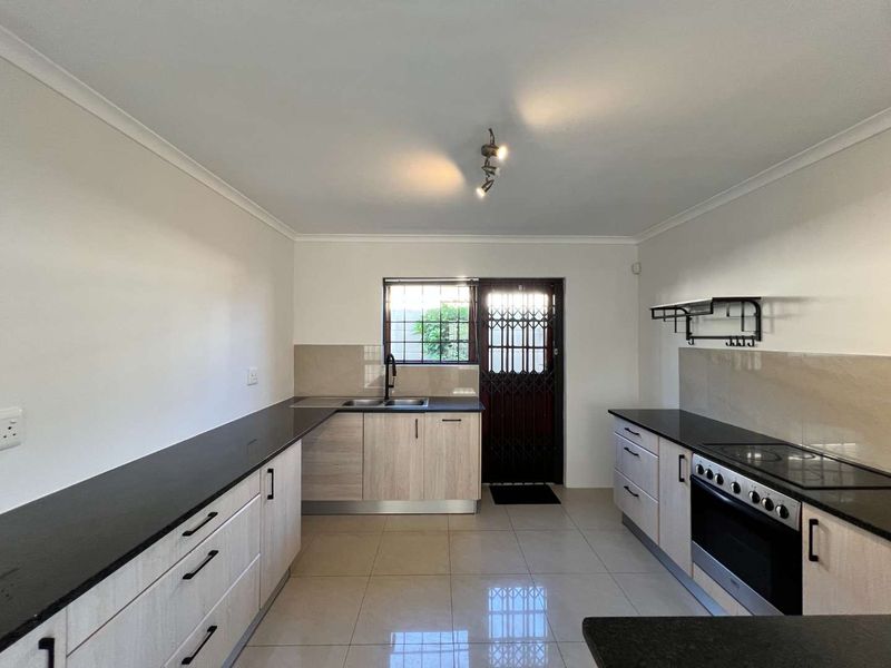 A clean, modern kitchen featuring light wood-finish cabinetry, dark stone countertops, integrated electric stove, and tiled flooring with direct access to the exterior.