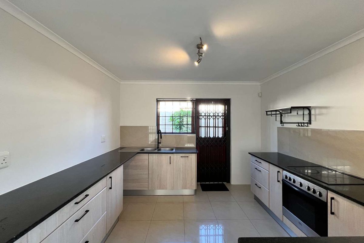 A clean, modern kitchen featuring light wood-finish cabinetry, dark stone countertops, integrated electric stove, and tiled flooring with direct access to the exterior.