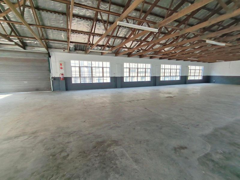 Spacious industrial warehouse unit featuring exposed timber roof trusses, concrete flooring, large steel-framed windows, and a roller shutter door for easy access.
