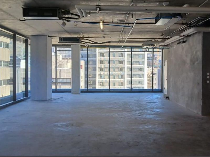An open-plan office space featuring industrial-style exposed concrete ceilings, ducting, and floor-to-ceiling windows with urban views.
