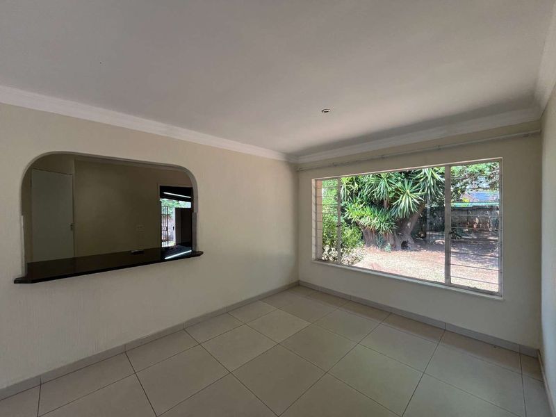 A bright, empty living room featuring large windows, tiled flooring, and a convenient serving hatch connecting to the adjacent kitchen area.